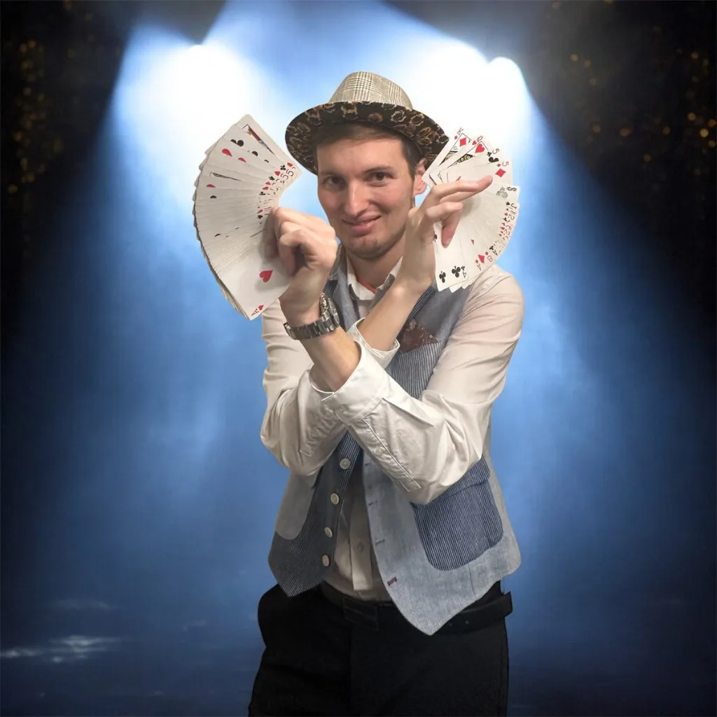 Kévin Boutillier en costume et chapeau, deux éventails de cartes, ambiance de scène et projecteurs